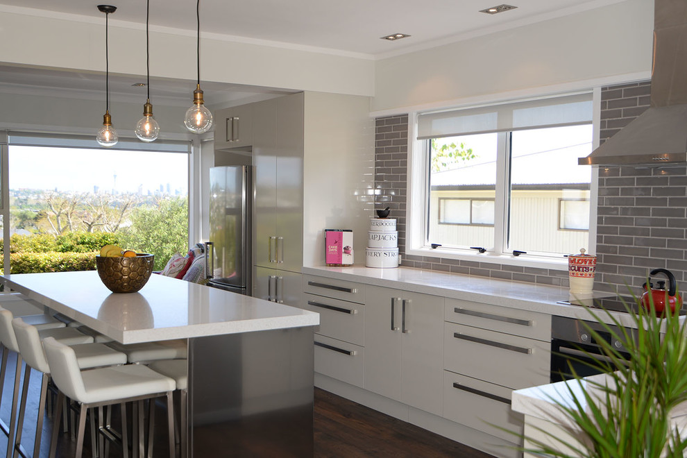 This is an example of a large modern l-shaped open plan kitchen in Auckland with an undermount sink, flat-panel cabinets, white cabinets, quartz benchtops, grey splashback, ceramic splashback, stainless steel appliances, dark hardwood floors, with island and brown floor.