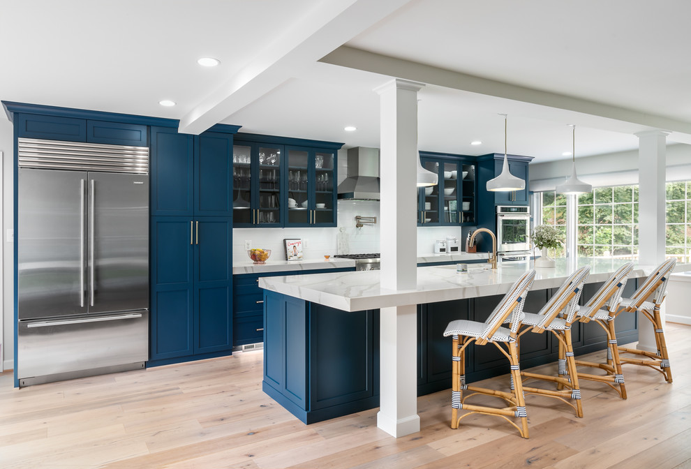 Photo of an expansive transitional galley open plan kitchen in DC Metro with an undermount sink, glass-front cabinets, blue cabinets, quartz benchtops, white splashback, ceramic splashback, stainless steel appliances, light hardwood floors, with island, beige floor and white benchtop.