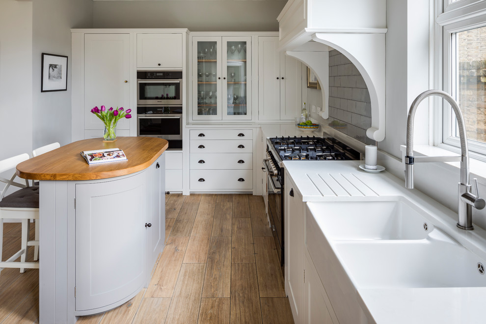 Inspiration for a country l-shaped kitchen in London with a farmhouse sink, shaker cabinets, white cabinets, wood benchtops, grey splashback, subway tile splashback, black appliances, medium hardwood floors, with island and brown floor.