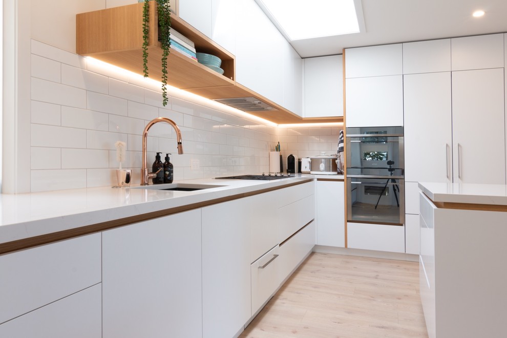 Photo of a mid-sized contemporary l-shaped open plan kitchen in Auckland with an undermount sink, recessed-panel cabinets, white cabinets, quartz benchtops, white splashback, porcelain splashback, panelled appliances, light hardwood floors, with island, beige floor and white benchtop.