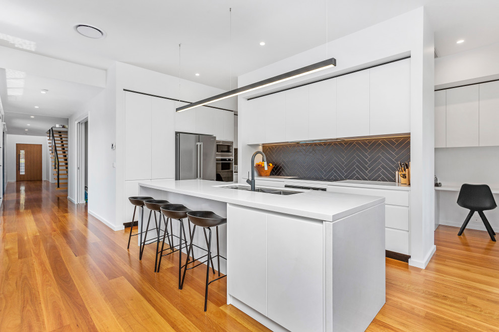 Contemporary l-shaped kitchen in Sydney with an undermount sink, flat-panel cabinets, white cabinets, grey splashback, stainless steel appliances, medium hardwood floors, with island, brown floor and white benchtop.