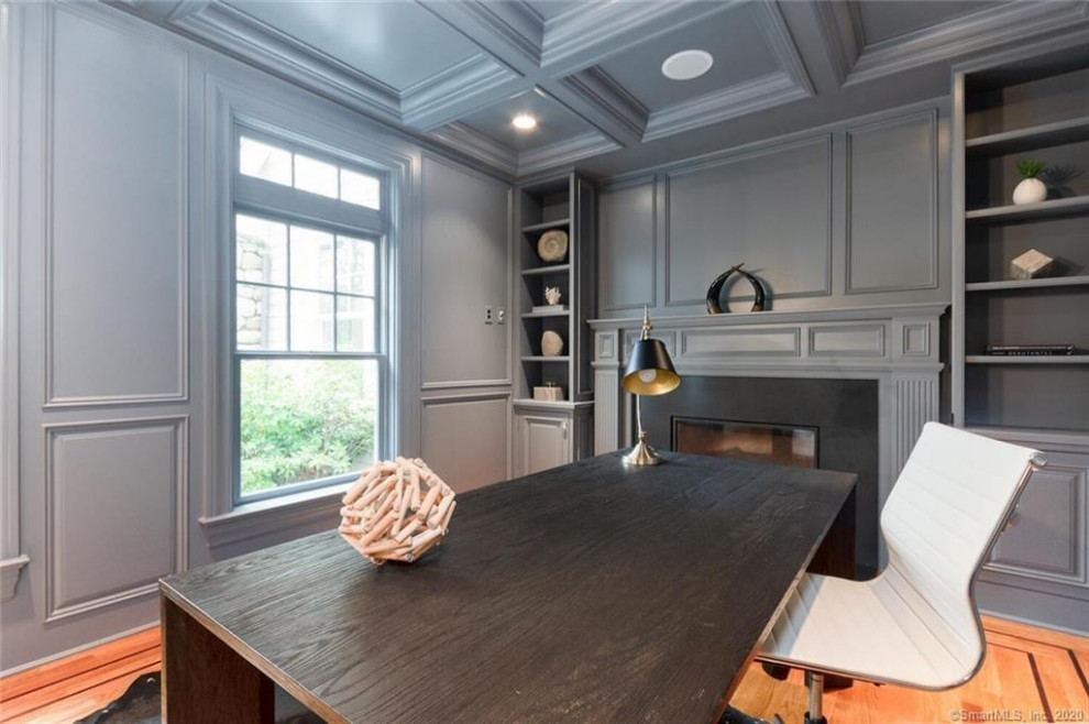 This is an example of a mid-sized modern home office in New York with grey walls, medium hardwood floors, a standard fireplace, a stone fireplace surround, a built-in desk, brown floor, coffered and panelled walls.