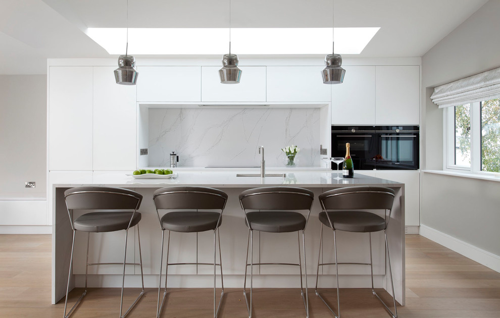 Photo of a mid-sized contemporary galley kitchen in Dublin with flat-panel cabinets, white splashback, marble splashback, black appliances, with island, an undermount sink, marble benchtops, light hardwood floors and beige floor.