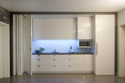 Concealed kitchen with white slab kitchen cabinets, bi-fold pocket doors, panel-ready tall cabinet (fridge), built-in microwave, and under-cabinet LED task lighting
