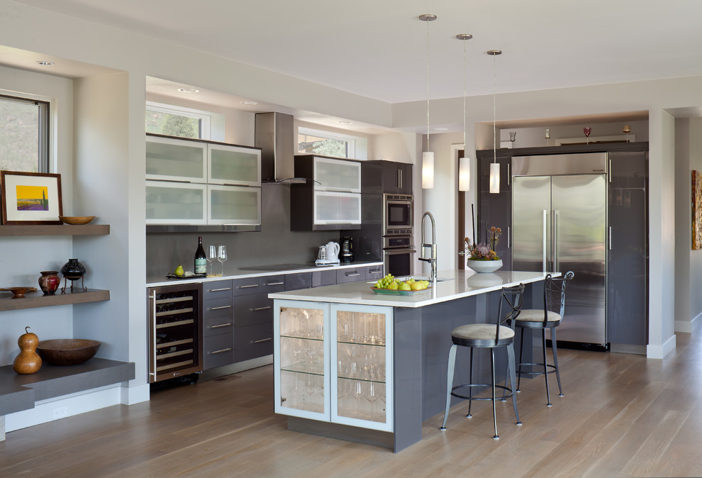 Design ideas for a large contemporary l-shaped open plan kitchen in Denver with flat-panel cabinets, grey cabinets, grey splashback, stainless steel appliances, light hardwood floors, with island, an undermount sink, glass sheet splashback, granite benchtops, beige floor and white benchtop.