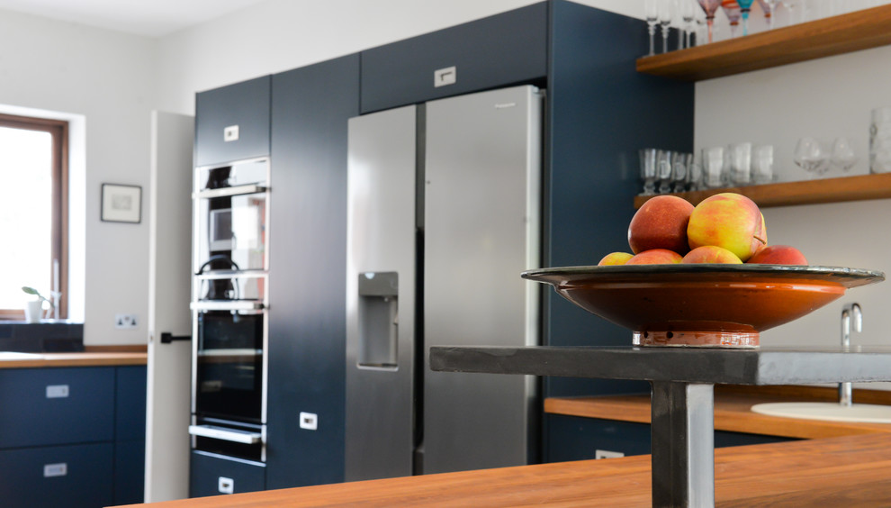 Mid-sized contemporary open plan kitchen in Sussex with a drop-in sink, flat-panel cabinets, blue cabinets, wood benchtops, black splashback, ceramic splashback, black appliances, concrete floors and with island.