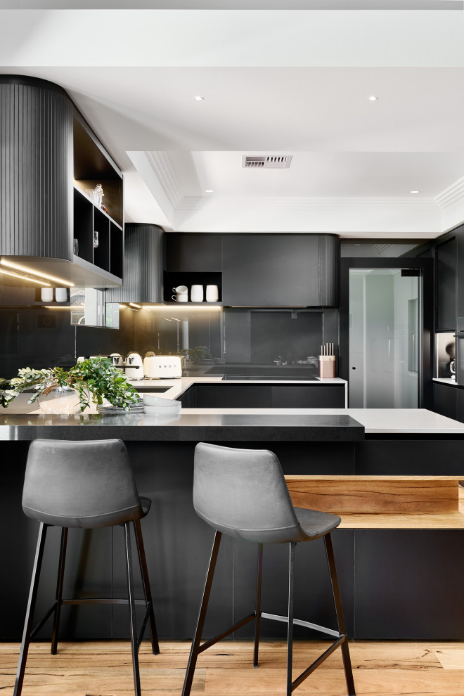 Inspiration for a large contemporary u-shaped open plan kitchen in Perth with an undermount sink, flat-panel cabinets, black cabinets, quartz benchtops, black splashback, glass sheet splashback, black appliances, light hardwood floors, with island, brown floor, grey benchtop and recessed.