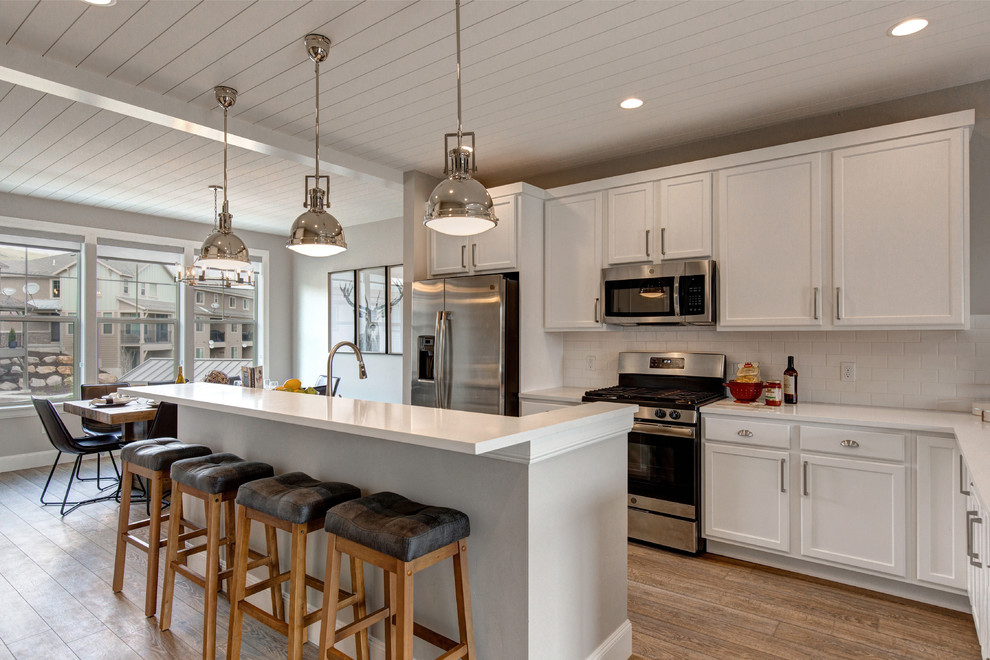 Example of a transitional l-shaped medium tone wood floor and brown floor kitchen design in Salt Lake City with shaker cabinets, white cabinets, white backsplash, subway tile backsplash, stainless steel appliances, an island and white countertops