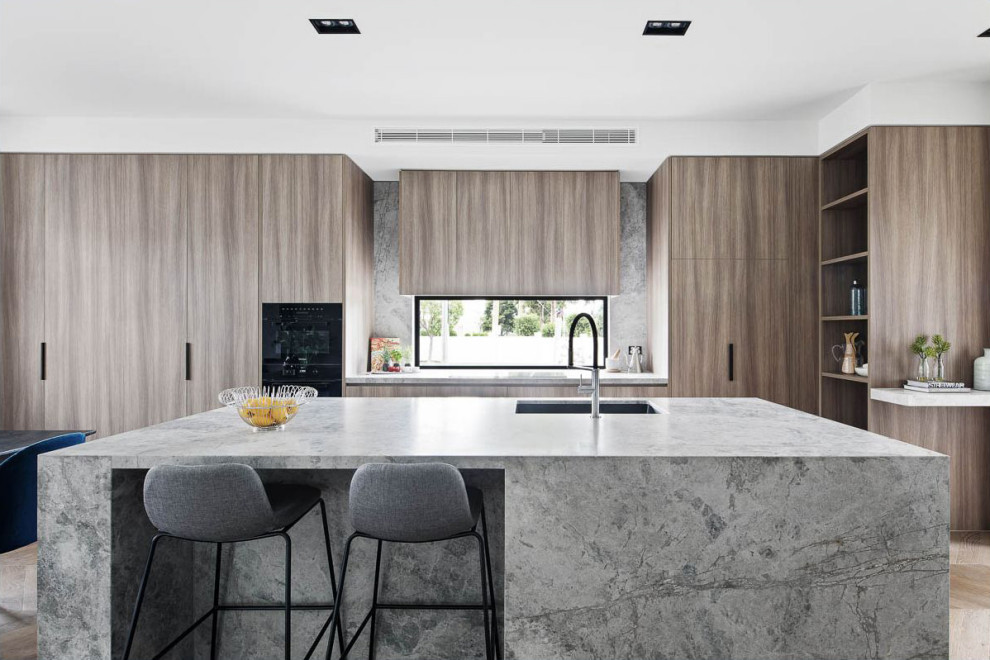 Photo of a mid-sized modern open plan kitchen in Melbourne with an undermount sink, light wood cabinets, marble benchtops, window splashback, black appliances, light hardwood floors, with island and grey benchtop.