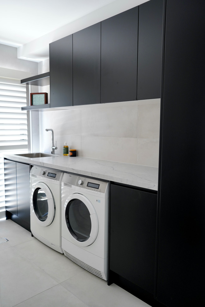 This is an example of a mid-sized modern single-wall dedicated laundry room in Sydney with a drop-in sink, flat-panel cabinets, black cabinets, quartz benchtops, grey splashback, cement tile splashback, white walls, a side-by-side washer and dryer and grey benchtop.