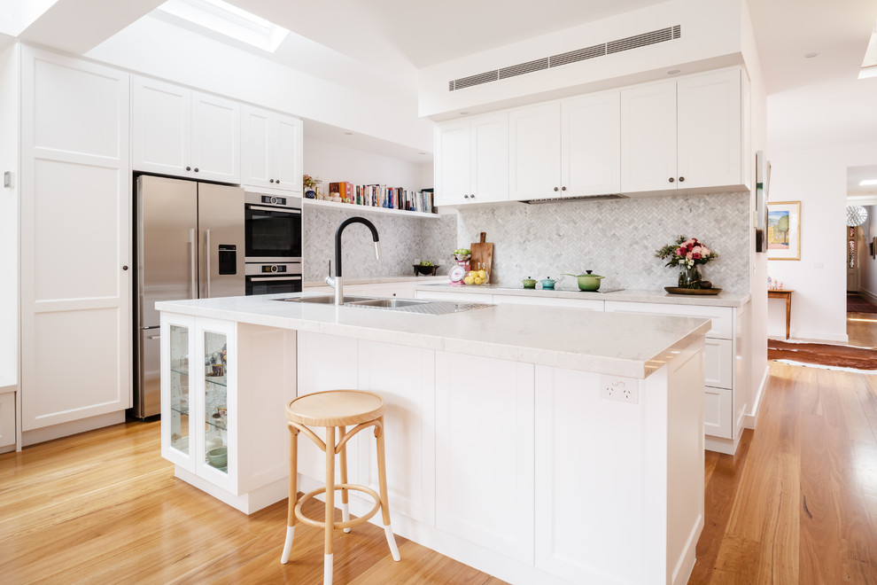 This is an example of a large transitional l-shaped open plan kitchen in Melbourne with a double-bowl sink, shaker cabinets, white cabinets, quartz benchtops, grey splashback, mosaic tile splashback, stainless steel appliances, light hardwood floors, with island and brown floor.