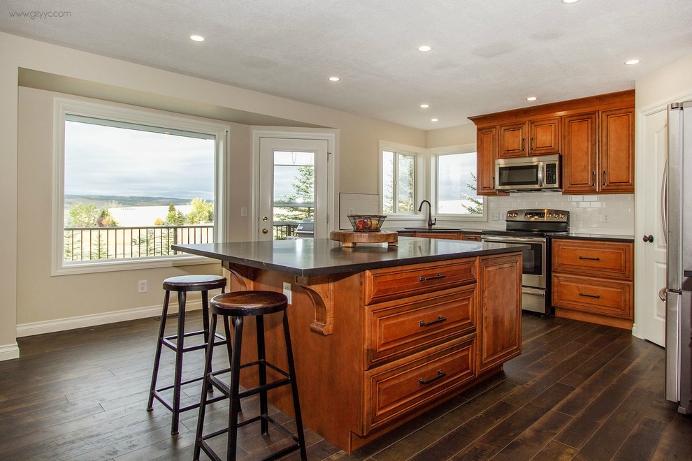 Example of a mid-sized mountain style l-shaped laminate floor and black floor eat-in kitchen design in Calgary with a drop-in sink, raised-panel cabinets, medium tone wood cabinets, quartzite countertops, white backsplash, ceramic backsplash, stainless steel appliances and an island