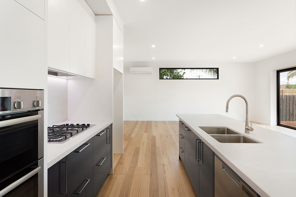 Photo of a mid-sized contemporary galley open plan kitchen in Melbourne with an undermount sink, flat-panel cabinets, white cabinets, solid surface benchtops, white splashback, ceramic splashback, stainless steel appliances, light hardwood floors, with island and white benchtop.