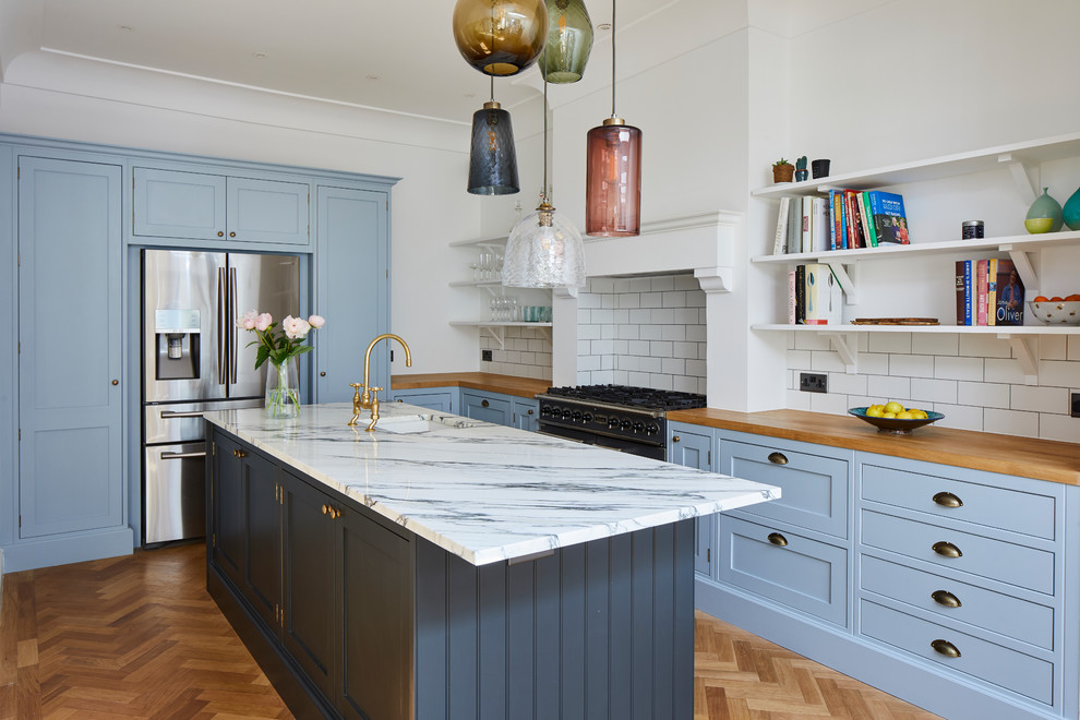 Transitional u-shaped kitchen in London with an undermount sink, shaker cabinets, blue cabinets, white splashback, subway tile splashback, stainless steel appliances, medium hardwood floors, with island, brown floor and brown benchtop.
