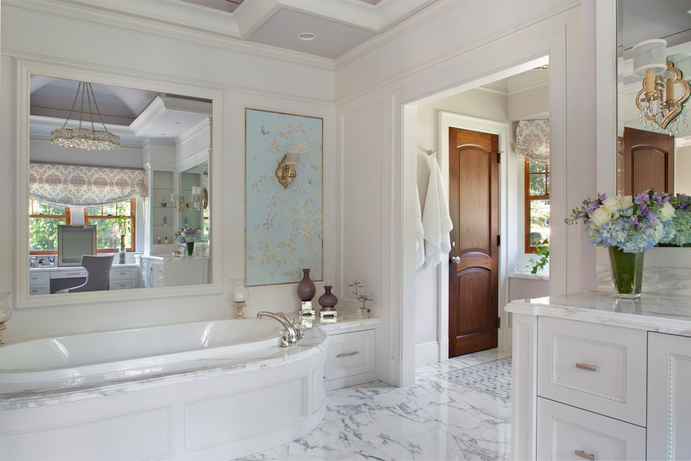 This is an example of a mid-sized transitional master bathroom in New York with recessed-panel cabinets, white cabinets, a drop-in tub, an alcove shower, a one-piece toilet, white tile, marble, white walls, marble floors, a drop-in sink, marble benchtops, white floor, a hinged shower door, white benchtops, an enclosed toilet, a double vanity, a built-in vanity, vaulted and decorative wall panelling.