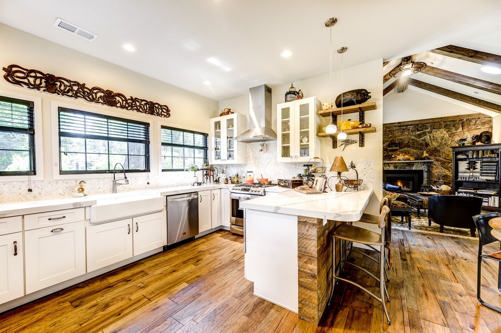Mid-sized country u-shaped open plan kitchen in Denver with a farmhouse sink, white cabinets, marble benchtops, white splashback, marble splashback, stainless steel appliances, medium hardwood floors, a peninsula, white benchtop and glass-front cabinets.