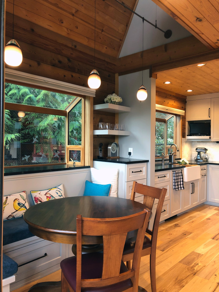 Medium sized farmhouse l-shaped kitchen/diner in Seattle with a belfast sink, shaker cabinets, grey cabinets, soapstone worktops, white splashback, marble splashback, stainless steel appliances, light hardwood flooring, yellow floors, black worktops and a vaulted ceiling.