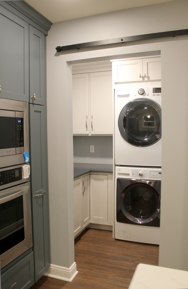 This is an example of a small transitional l-shaped dedicated laundry room in Other with shaker cabinets, quartz benchtops, white splashback, ceramic splashback, vinyl floors, brown floor, blue benchtop, grey cabinets, blue walls and a stacked washer and dryer.