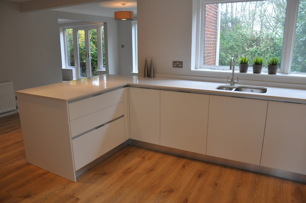 Small contemporary u-shaped eat-in kitchen in Cheshire with a drop-in sink, flat-panel cabinets, white cabinets, solid surface benchtops, white splashback, glass sheet splashback, stainless steel appliances, medium hardwood floors and a peninsula.