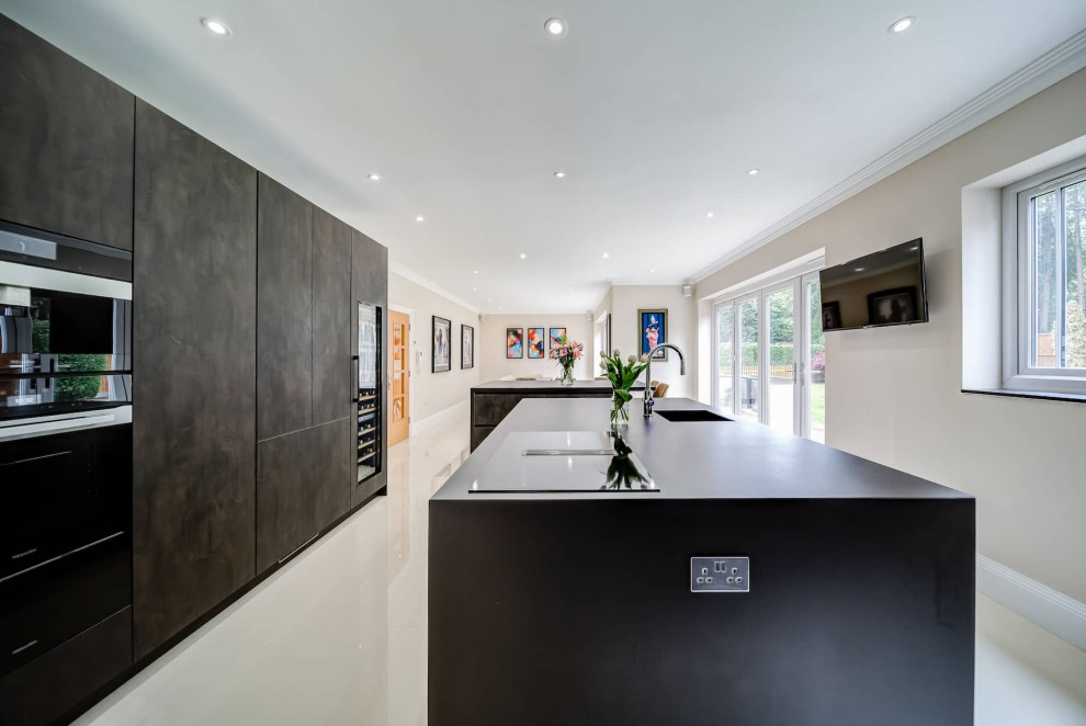 Large minimalist galley porcelain tile and white floor open concept kitchen photo in London with a drop-in sink, flat-panel cabinets, dark wood cabinets, black appliances, two islands and black countertops