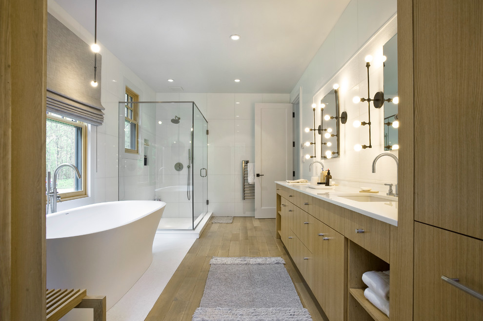 Photo of a transitional master bathroom in New York with flat-panel cabinets, light wood cabinets, a freestanding tub, a corner shower, a one-piece toilet, white tile, white walls, light hardwood floors, an undermount sink, engineered quartz benchtops, a hinged shower door and white benchtops.