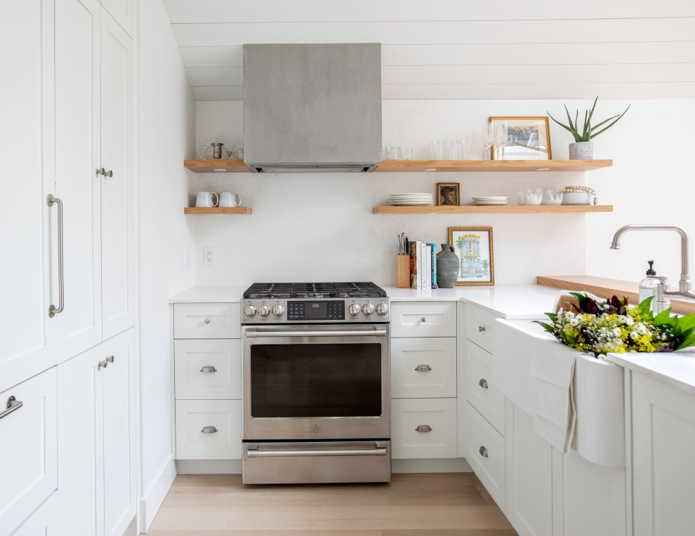 Photo of a beach style u-shaped kitchen in Vancouver with a farmhouse sink, shaker cabinets, white cabinets, stainless steel appliances, light hardwood floors, a peninsula, beige floor, white benchtop, timber and vaulted.