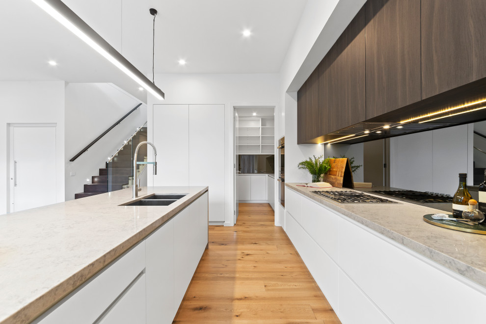 Photo of a large contemporary l-shaped eat-in kitchen in Melbourne with a drop-in sink, flat-panel cabinets, white cabinets, mirror splashback, stainless steel appliances, medium hardwood floors, with island, brown floor and white benchtop.