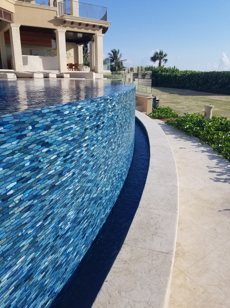 This is an example of a large mediterranean backyard custom-shaped infinity pool in Miami with a water feature and natural stone pavers.