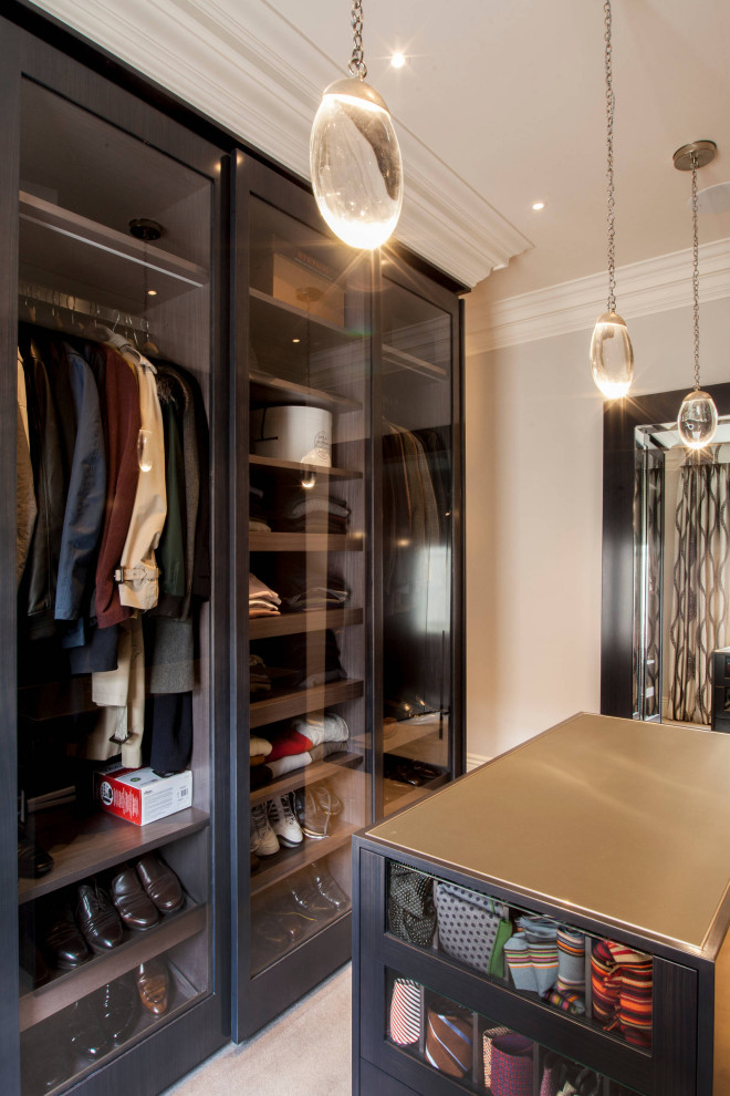 This is an example of a mid-sized contemporary men's walk-in wardrobe in London with glass-front cabinets, dark wood cabinets, carpet and beige floor.