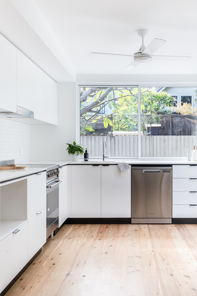 This is an example of a small contemporary l-shaped eat-in kitchen in Sydney with an undermount sink, flat-panel cabinets, white cabinets, quartz benchtops, white splashback, mosaic tile splashback, stainless steel appliances, medium hardwood floors, no island and white benchtop.