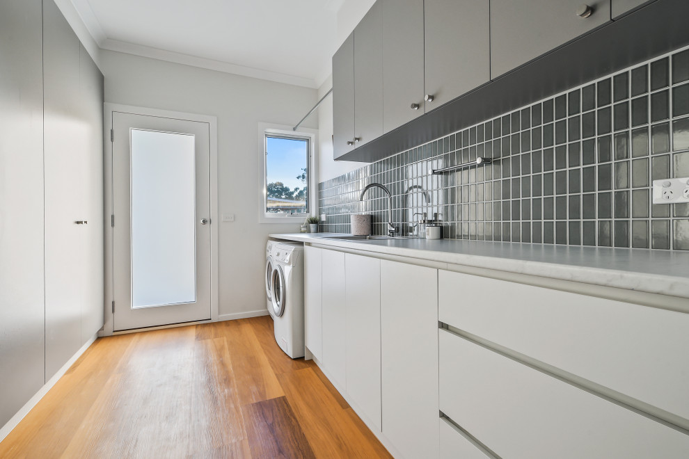 This is an example of a mid-sized country galley utility room in Other with a single-bowl sink, white cabinets, laminate benchtops, green splashback, ceramic splashback, white walls, vinyl floors, a side-by-side washer and dryer, brown floor and white benchtop.