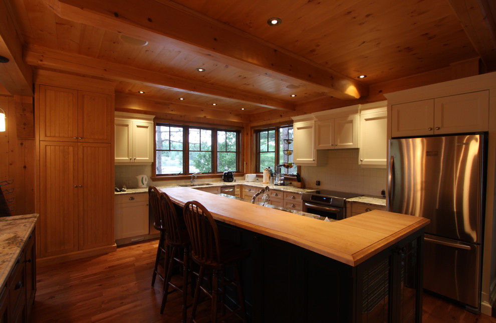Photo of a mid-sized country l-shaped eat-in kitchen in Toronto with an undermount sink, recessed-panel cabinets, white cabinets, wood benchtops, white splashback, ceramic splashback, stainless steel appliances, medium hardwood floors and with island.
