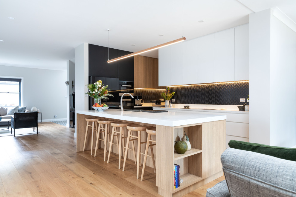 Design ideas for a large contemporary kitchen pantry in Sydney with a drop-in sink, flat-panel cabinets, quartz benchtops, black splashback, mosaic tile splashback, black appliances, light hardwood floors, with island and white benchtop.