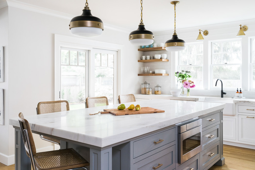 This is an example of a large traditional l-shaped open plan kitchen in Boston with a farmhouse sink, shaker cabinets, white cabinets, marble benchtops, stainless steel appliances, with island, light hardwood floors, white splashback and subway tile splashback.
