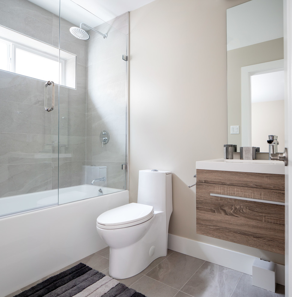 Inspiration for a small contemporary 3/4 bathroom in Vancouver with a one-piece toilet, laminate floors, a console sink, glass benchtops, brown floor, blue benchtops, flat-panel cabinets, brown cabinets, an undermount tub, a shower/bathtub combo, gray tile, porcelain tile, beige walls and a hinged shower door.