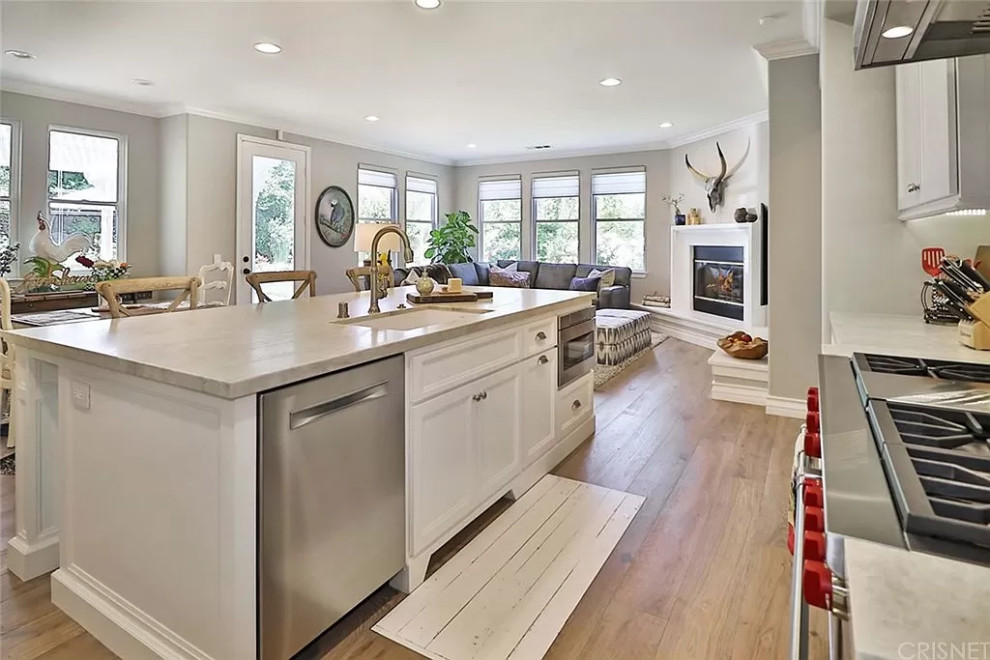 Design ideas for a large country l-shaped open plan kitchen in Los Angeles with an undermount sink, shaker cabinets, white cabinets, quartzite benchtops, white splashback, porcelain splashback, stainless steel appliances, medium hardwood floors, with island, brown floor and beige benchtop.