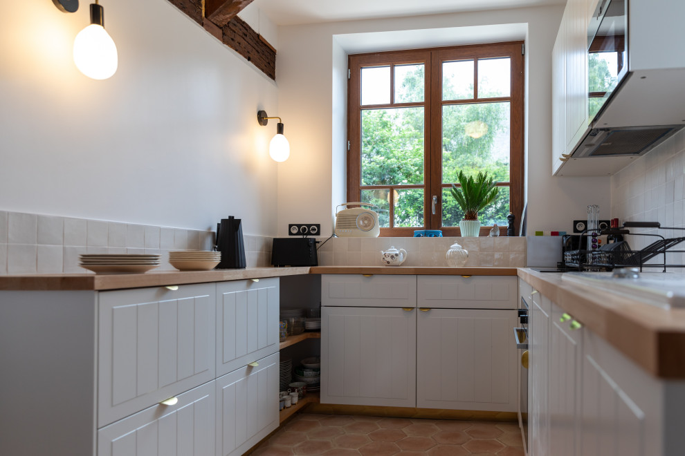 Photo of a mid-sized country u-shaped open plan kitchen in Le Havre with an undermount sink, beaded inset cabinets, white cabinets, wood benchtops, white splashback, panelled appliances, terra-cotta floors, no island, pink floor, beige benchtop and exposed beam.