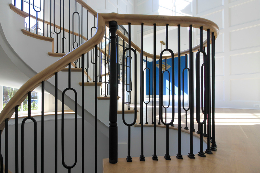 На фото: огромная изогнутая деревянная лестница в стиле неоклассика (современная классика) с деревянными ступенями, перилами из смешанных материалов и панелями на стенах с