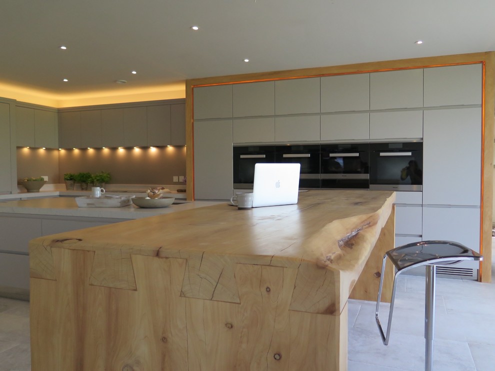 This is an example of an expansive contemporary l-shaped open plan kitchen in West Midlands with a double-bowl sink, flat-panel cabinets, grey cabinets, solid surface benchtops, green splashback, glass sheet splashback, black appliances, ceramic floors and a peninsula.