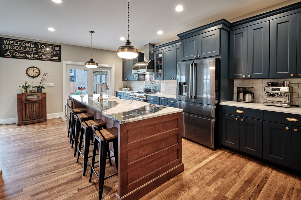 This is an example of a large eclectic galley open plan kitchen in Philadelphia with a farmhouse sink, recessed-panel cabinets, blue cabinets, quartzite benchtops, grey splashback, ceramic splashback, black appliances, medium hardwood floors, with island, brown floor and multi-coloured benchtop.
