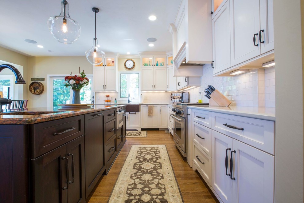 Large transitional l-shaped light wood floor and brown floor enclosed kitchen photo with a farmhouse sink, shaker cabinets, white cabinets, solid surface countertops, white backsplash, porcelain backsplash, stainless steel appliances and an island