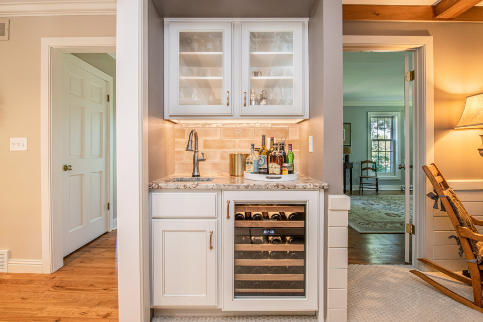 This is an example of a large traditional u-shaped home bar in Milwaukee with an undermount sink, recessed-panel cabinets, white cabinets, granite benchtops, beige splashback, subway tile splashback, medium hardwood floors, multi-coloured floor and beige benchtop.