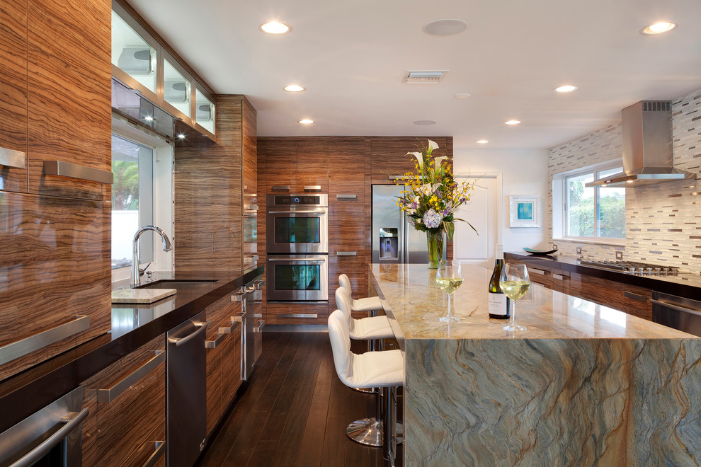 Photo of an expansive contemporary galley separate kitchen in Miami with an undermount sink, flat-panel cabinets, medium wood cabinets, multi-coloured splashback, matchstick tile splashback, stainless steel appliances, dark hardwood floors, with island, brown floor and black benchtop.