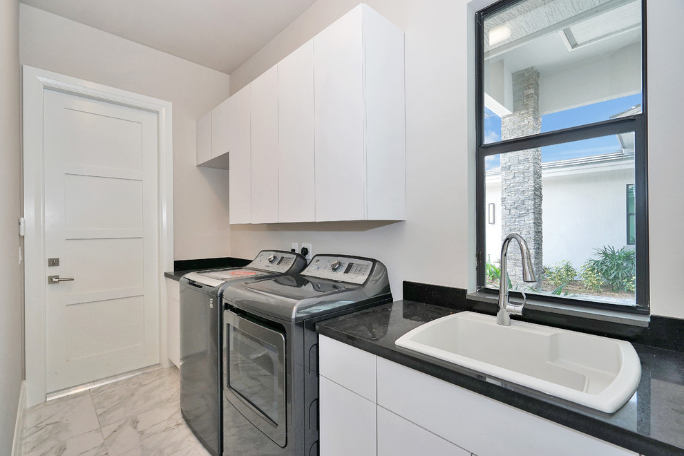 Design ideas for a mid-sized contemporary single-wall dedicated laundry room in Miami with a drop-in sink, flat-panel cabinets, white cabinets, granite benchtops, beige walls, marble floors, a side-by-side washer and dryer, white floor and black benchtop.