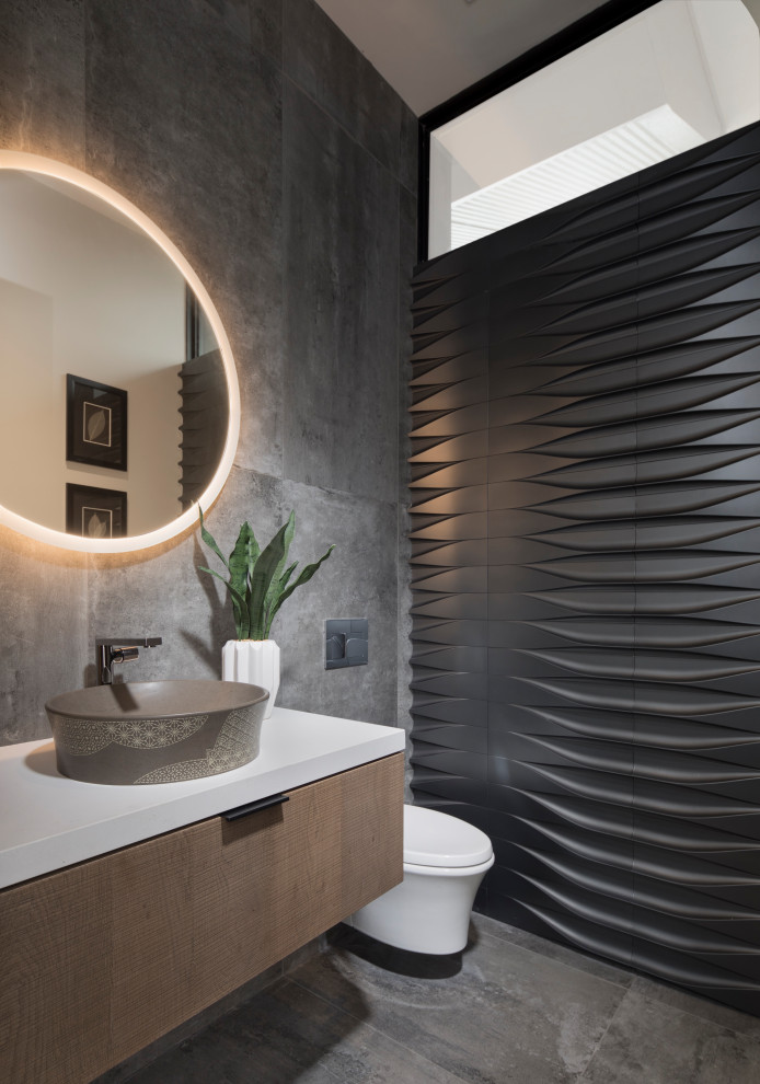 Inspiration for a mid-sized contemporary powder room in Las Vegas with flat-panel cabinets, medium wood cabinets, a wall-mount toilet, porcelain tile, porcelain floors, a vessel sink, engineered quartz benchtops, grey floor, white benchtops, gray tile and grey walls.