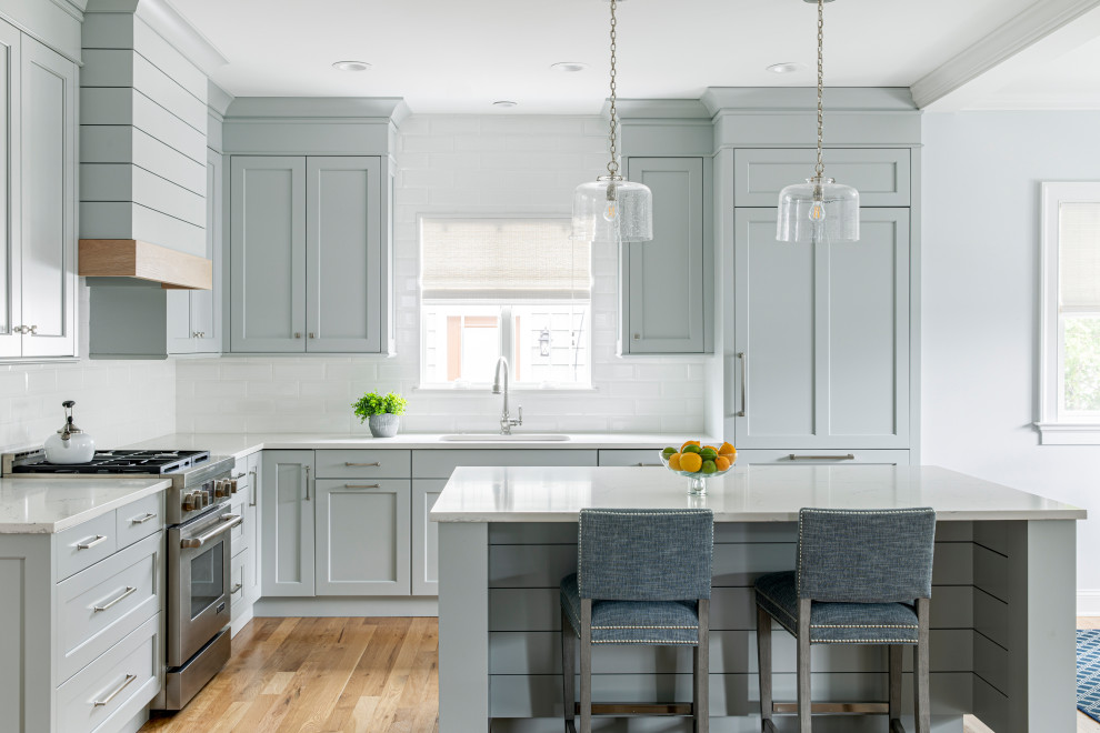 Beach style l-shaped kitchen in New York with an undermount sink, shaker cabinets, blue cabinets, quartz benchtops, white splashback, subway tile splashback, panelled appliances, light hardwood floors, with island and white benchtop.