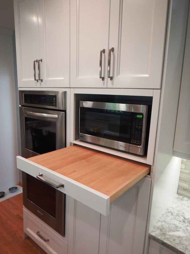 Example of a large transitional l-shaped light wood floor eat-in kitchen design with an undermount sink, recessed-panel cabinets, white cabinets, quartz countertops, gray backsplash, stone tile backsplash, stainless steel appliances and an island