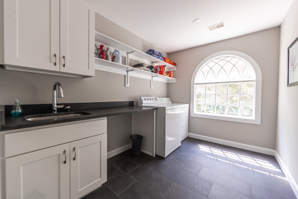 This is an example of a mid-sized traditional single-wall dedicated laundry room in Baltimore with an undermount sink, shaker cabinets, white cabinets, solid surface benchtops, grey walls, slate floors, a side-by-side washer and dryer, black floor and black benchtop.
