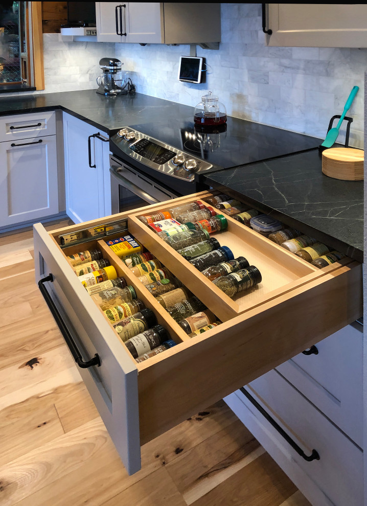 Photo of a mid-sized country l-shaped eat-in kitchen in Seattle with a farmhouse sink, shaker cabinets, grey cabinets, soapstone benchtops, white splashback, marble splashback, stainless steel appliances, light hardwood floors, yellow floor, black benchtop and vaulted.