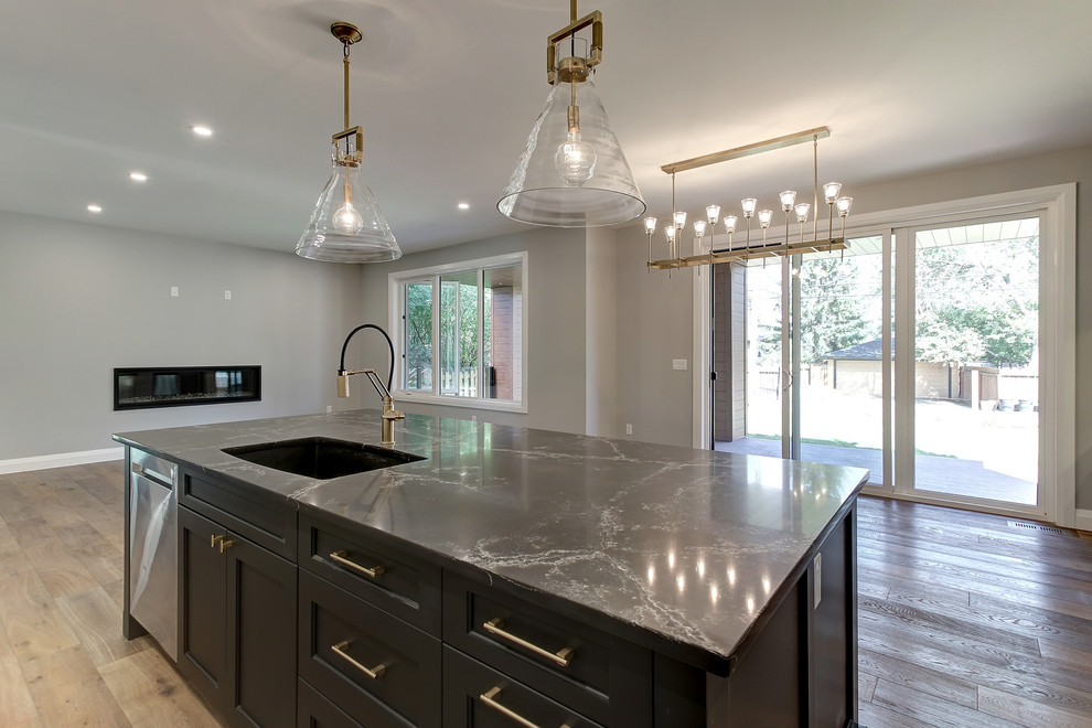 Open concept kitchen - mid-sized transitional l-shaped medium tone wood floor and brown floor open concept kitchen idea in Edmonton with an undermount sink, recessed-panel cabinets, white cabinets, quartz countertops, white backsplash, ceramic backsplash, stainless steel appliances, an island and black countertops